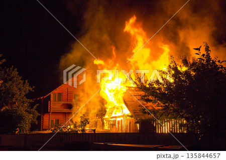 Burning wooden house at night. Bright orange flames and dense smoke from under the tiled roof on dark sky, trees silhouettes and residential neighbor cottage background. Disaster and danger concept. Burning wooden house at night. Bright orange flames and dense smoke from under the tiled roof on dark sky, trees silhouettes and residential neighbor cottage background. Disaster and danger concept. 135844657