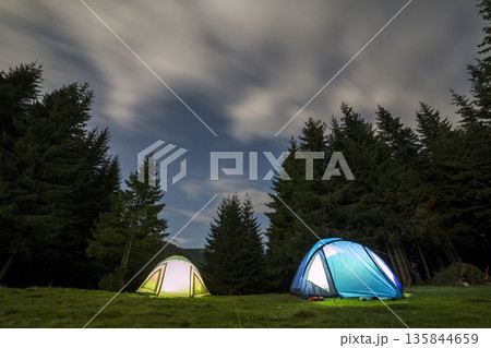 Bright big moon in dark blue cloudy sky over two tourist tents on green grassy forest clearing among tall pine trees on distant mountain background. Tourism, night camping in summer mountains. Bright big moon in dark blue cloudy sky over two tourist tents on green grassy forest clearing among tall pine trees on distant mountain background. Tourism, night camping in summer mountains. 135844659