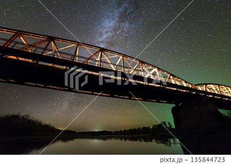 Illuminated metal bridge on concrete supports reflected in water on dark starry sky with Milky Way constellation background. Night photography concept. Illuminated metal bridge on concrete supports reflected in water on dark starry sky with Milky Way constellation background. Night photography concept. 135844723