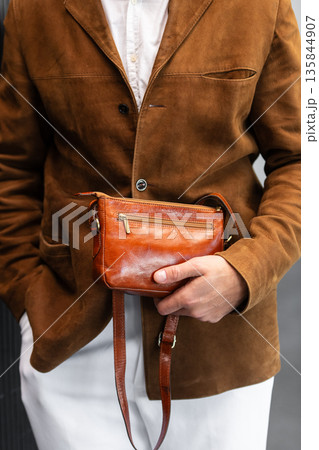Close up of a stylish man in a brown suede jacket holding a tan leather wristlet clutch bag Close up of a stylish man in a brown suede jacket holding a tan leather wristlet clutch bag 135844907