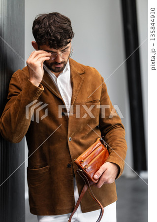 Close up of a stylish man in a brown suede jacket holding a tan leather wristlet clutch bag Close up of a stylish man in a brown suede jacket holding a tan leather wristlet clutch bag 135844910