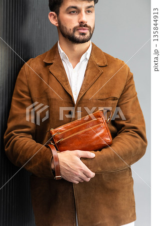 Close up of a stylish man in a brown suede jacket holding a tan leather wristlet clutch bag Close up of a stylish man in a brown suede jacket holding a tan leather wristlet clutch bag 135844913