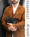 Stylish man in a brown suede blazer and white trousers leaning against a wall holding a small dark leather crossbody bag 135844916