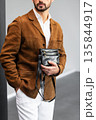 Stylish man in a brown suede blazer and white trousers leaning against a wall holding a small dark leather crossbody bag 135844917