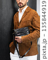 Stylish man in a brown suede blazer and white trousers leaning against a wall holding a small dark leather crossbody bag 135844919