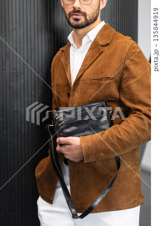 Stylish man in a brown suede blazer and white trousers leaning against a wall holding a small dark leather crossbody bag Stylish man in a brown suede blazer and white trousers leaning against a wall holding a small dark leather crossbody bag 135844919