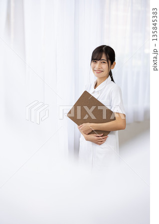 バインダーを抱えて微笑む白衣を着た若い女性 135845383