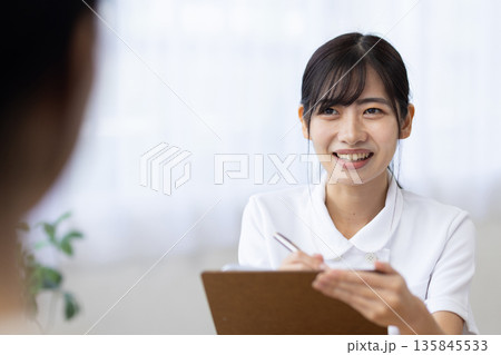 問診表に記入しながら患者と話す白衣を着た笑顔の若い女性 問診表に記入しながら患者と話す白衣を着た笑顔の若い女性 135845533