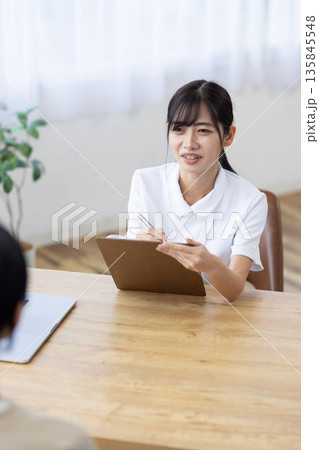 問診表に記入しながら患者と話す白衣を着た笑顔の若い女性 135845548