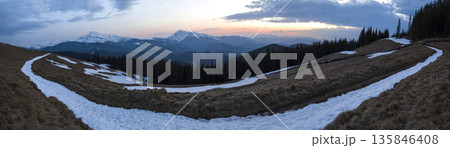 Panorama of beautiful spring sunset in Carpathian mountains. Valley with dry grass and snow, transparent fresh air, dense evergreen forest and soft sun glow over distant snow-covered mountain range. Panorama of beautiful spring sunset in Carpathian mountains. Valley with dry grass and snow, transparent fresh air, dense evergreen forest and soft sun glow over distant snow-covered mountain range. 135846408