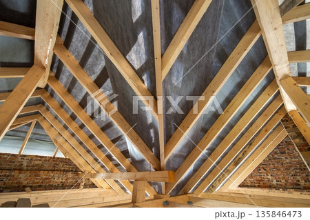 Attic of a building under construction with wooden beams of a roof structure and brick walls. 135846473