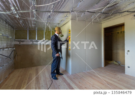 Strange man in businessman suit and gas protection mask inside a room under renovation works holding electric screwdriver and a level tools. 135846479
