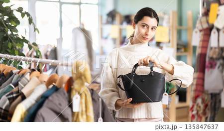 Fashionable young girl customer standing near shelves with clothes in store and choosing handbag 135847637