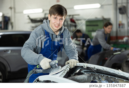 Male mechanic prepares car for painting in workshop - using grinding machine he sands bumper before painting Male mechanic prepares car for painting in workshop - using grinding machine he sands bumper before painting 135847887