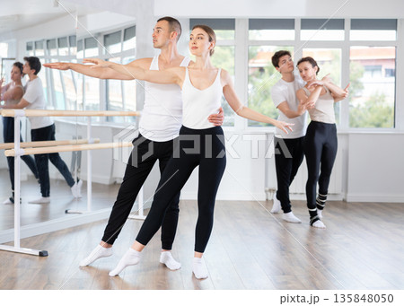 Smiling young man and female partner in black and white clothing with group active people training ballet dance movements in dance salon Smiling young man and female partner in black and white clothing with group active people training ballet dance movements in dance salon 135848050