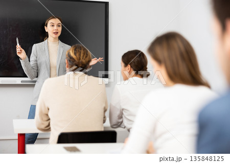 Positive female trainer standing near interactive board and conducting advanced training courses to office employees sitting at desks in classroom Positive female trainer standing near interactive board and conducting advanced training courses to office employees sitting at desks in classroom 135848125