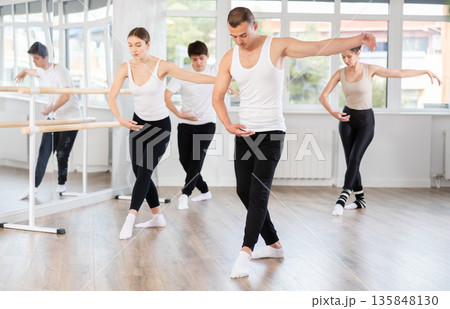 Experienced young male ballet dancer, dressed in white and black executing dance moves with focused precision among group of people rehearsing in ballet studio Experienced young male ballet dancer, dressed in white and black executing dance moves with focused precision among group of people rehearsing in ballet studio 135848130