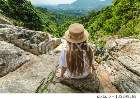 Young woman sitting on rocky ledge at Na Muang Waterfall 2 viewpoint in Koh Samui Thailand, overlooking lush tropical valley and distant mountains under bright sky 135849569