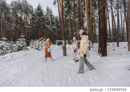 Two teenagers and adult woman walking through snowy pine forest during winter outdoor family walk and peaceful nature escape 135849592