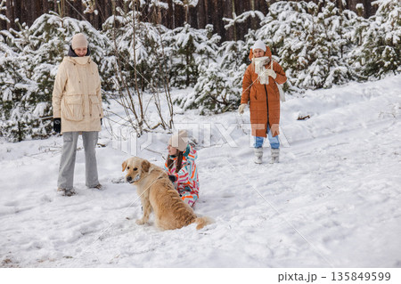 Teenage girl playing with Golden Retriever dog on snowy forest trail while two women walk behind, winter outdoor family leisure, pet companionship, calm seasonal lifestyle scene Teenage girl playing with Golden Retriever dog on snowy forest trail while two women walk behind, winter outdoor family leisure, pet companionship, calm seasonal lifestyle scene 135849599