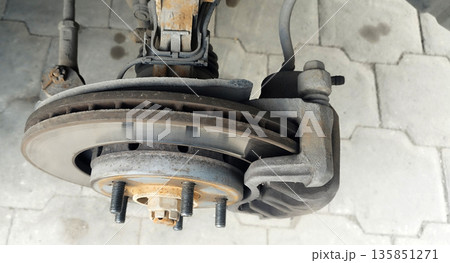 Close-up of a dirty, ventilated disc brake assembly with a caliper showing wear marks on the rotor and rusty wheel studs against a light grey asphalt background 135851271