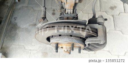 Close-up of a dirty, vented disc brake assembly with caliper, showing visible wear marks on the rotor and rusty wheel studs against a light gray paved background 135851272
