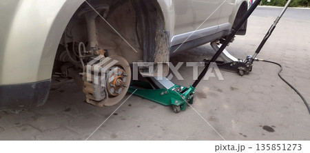 The beige car is lifted with a green jack, revealing a dusty rear disc brake, suggesting the wheel has been removed for repair or outdoor maintenance. 135851273