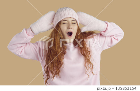 Funny woman in pink winter hat, mittens and sweatshirt stretching and yawning on beige background 135852154