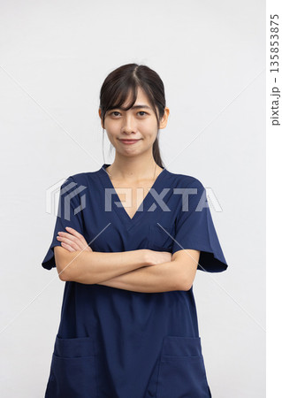 白背景の前で腕組みをして微笑む紺色の医療用スクラブを着た若い女性 白背景の前で腕組みをして微笑む紺色の医療用スクラブを着た若い女性 135853875