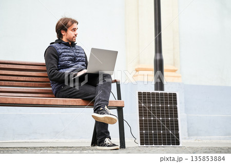 Man sits on wooden bench, working on laptop connected to solar panel. Urban setting and casual attire highlight modern, sustainable, mobile, eco-friendly workspace. Man sits on wooden bench, working on laptop connected to solar panel. Urban setting and casual attire highlight modern, sustainable, mobile, eco-friendly workspace. 135853884