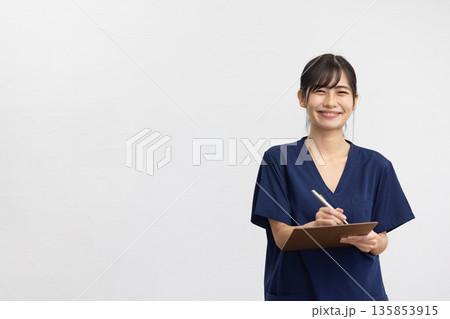 白背景の前でバインダーとペンを持ち微笑む紺色のスクラブを着た若い女性 白背景の前でバインダーとペンを持ち微笑む紺色のスクラブを着た若い女性 135853915