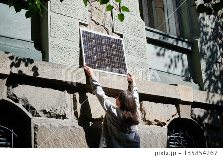 Woman holding photovoltaic solar panel in front of historical building, wearing plaid shirt and sunglasses. Concept of integration of sustainable renewable energy sources into old architecture. 135854267