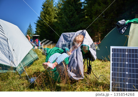 Young child plays with smartphone while charging with photovoltaic solar panel near tourist tent in summer. Integration of renewable energy in outdoor camping activities. 135854268