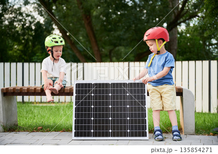 Two young boys sit on wooden bench near solar panel in park. Children in helmets, having rest after riding balance bike. Background features white fence and trees. Concept of green energy generation. 135854271