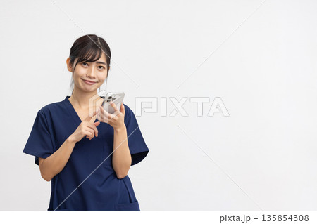 白背景の前でスマートフォンを手にカメラ目線で微笑む紺色のスクラブを着た若い女性 白背景の前でスマートフォンを手にカメラ目線で微笑む紺色のスクラブを着た若い女性 135854308