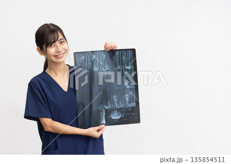 白背景の前でレントゲン写真を手に微笑む紺色のスクラブを着た若い女性 135854511