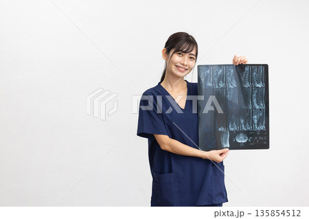 白背景の前でレントゲン写真を手に微笑む紺色のスクラブを着た若い女性 白背景の前でレントゲン写真を手に微笑む紺色のスクラブを着た若い女性 135854512