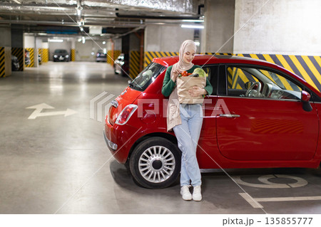 A woman wearing a hijab stands in a parking garage with a bag of fresh groceries next to her red car A woman wearing a hijab stands in a parking garage with a bag of fresh groceries next to her red car 135855777