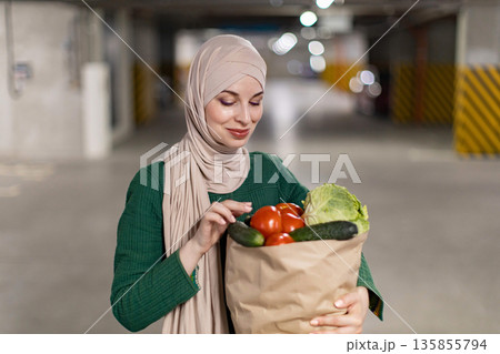 A woman wearing a hijab carries a bag of fresh produce in a parking garage, likely after shopping A woman wearing a hijab carries a bag of fresh produce in a parking garage, likely after shopping 135855794