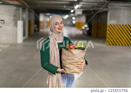 A smiling woman in a hijab carries a bag of fresh produce in a parking garage, ready to head home 135855798