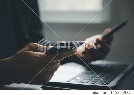 Close-up of a hand holding a credit card, with a blurred background of a laptop and another hand holding a phone. Close-up of a hand holding a credit card, with a blurred background of a laptop and another hand holding a phone. 135855987