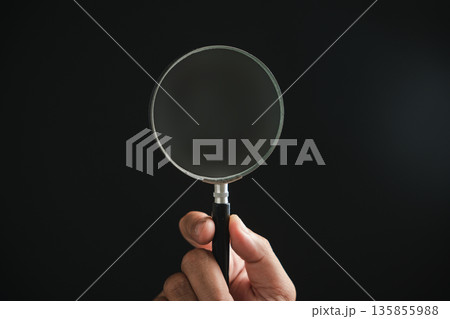 Hand holding a magnifying glass against a black background. The glass is slightly dusty. 135855988