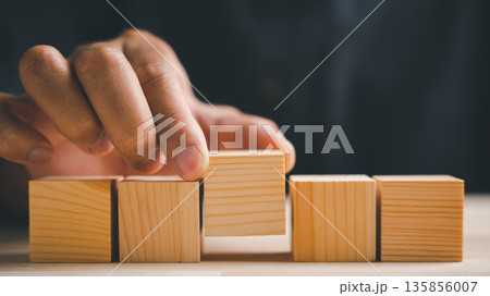 A hand places a wooden block on top of other blocks, forming a line. The blocks are made of natural wood. A hand places a wooden block on top of other blocks, forming a line. The blocks are made of natural wood. 135856007