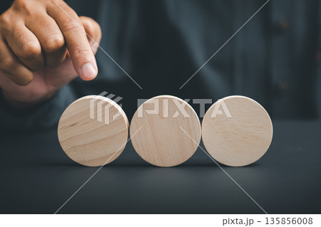 Three wooden circles on a dark surface, a hand points at the middle circle. 135856008