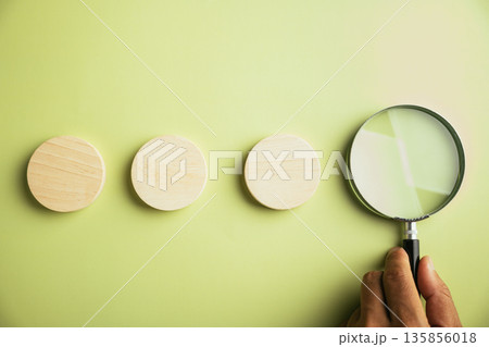 Three wooden circles arranged in a row, with a magnifying glass positioned to the right. A hand holds the magnifying glass. The background is a light green. Three wooden circles arranged in a row, with a magnifying glass positioned to the right. A hand holds the magnifying glass. The background is a light green. 135856018