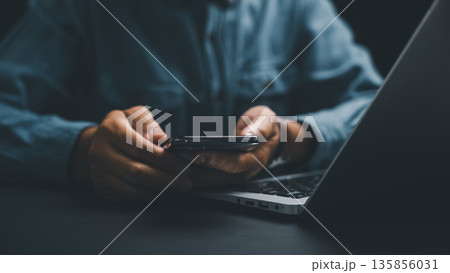 Close-up of hands holding a smartphone, blurred laptop in the background. Dark, moody aesthetic. Close-up of hands holding a smartphone, blurred laptop in the background. Dark, moody aesthetic. 135856031