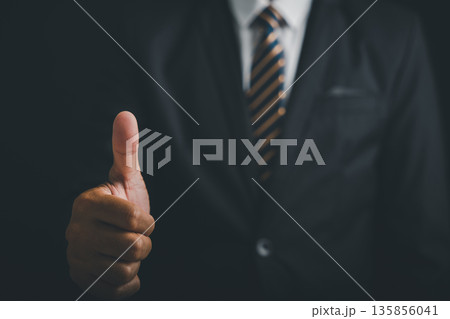 Close-up of a man's hand giving a thumbs up, blurred suit jacket and striped tie in the background. 135856041