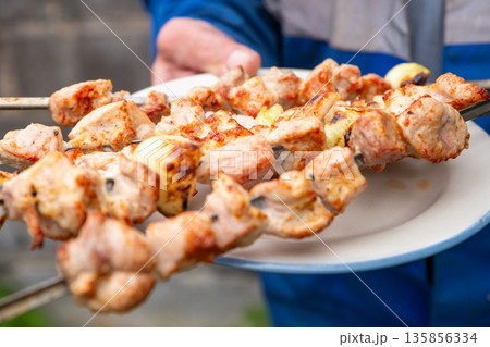 Man holding skewers of roasted pork shashlik over a plate. Traditional eastern european grilled meat dish for picnic, outdoor cooking food. 135856334