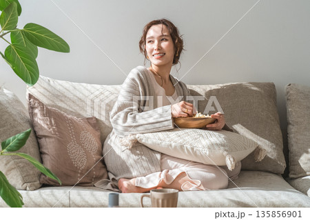 リビングのソファに座りポップコーンを食べる笑顔のアジア人女性 リビングのソファに座りポップコーンを食べる笑顔のアジア人女性 135856901