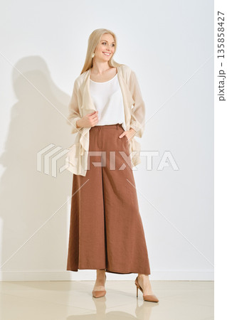 A smiling woman stands in a bright studio dressed in a light top and wide-leg brown pants. 135858427